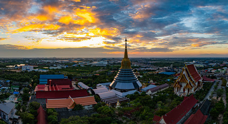 An aerial Wat Ketmadi Sriwararam perspective showcasing a stunning Thai temple complex with intricate golden spires, surrounded by urban and rural landscapes under a cloudy blue sky.の写真素材