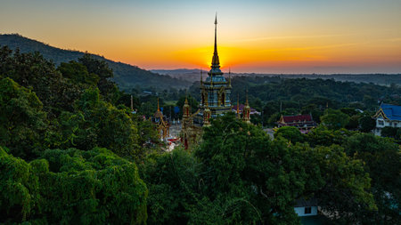 A breathtaking aerial view of a golden Buddhist temple at Wat Mae Klang Waterfall in Chiang Mai Thailand is surrounded by lush greenery, with the sunrise casting warm hues across the horizon.の写真素材