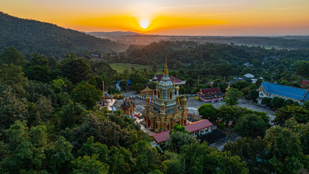A breathtaking aerial view of a golden Buddhist temple at Wat Mae Klang Waterfall in Chiang Mai Thailand is surrounded by lush greenery, with the sunrise casting warm hues across the horizon.の写真素材