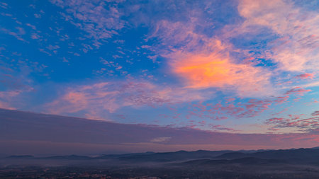 Aerial shot of a gorgeous sunrise. A panoramic view of dramatic mountain peaks silhouetted against a vibrant sunset sky, with fiery hues of red, orange, and pink blending into the fading blue.の写真素材