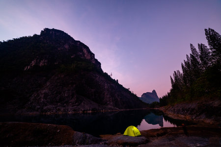 Serene wilderness view of a lone camper near a tent by a tranquil mountain lake at sunrise, with rugged cliffs and forests creating a breathtaking natural landscape.の写真素材