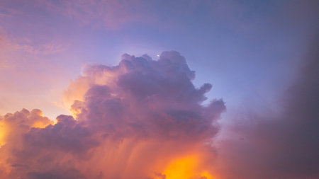 A vivid and dynamic sky captures towering storm clouds illuminated by the warm hues of a setting sun, creating a striking contrast between light and shadow above a darkened landscape.の写真素材