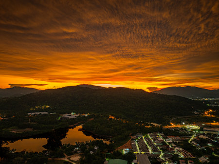 A breathtaking aerial view of a vivid orange sunset casting dramatic light over forested hills and a reflective river, with urban elements nestled at the landscapeâs edge beneath a glowing sky.の写真素材
