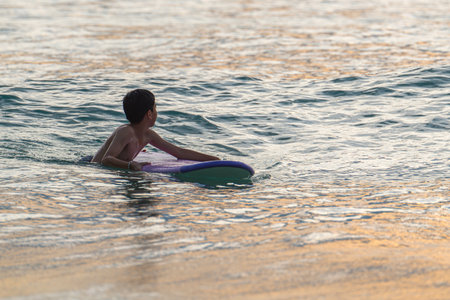 A young surfer rides with confidence and excitement, enjoying the calm waves of a tropical beach. The warm hues of sunset reflect on the ocean, blending energy, motion, and carefree joy.の写真素材