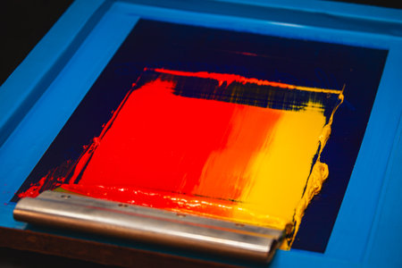 An artisan applies layers of warm colored inks on a screen, blending fiery hues to produce a smooth...