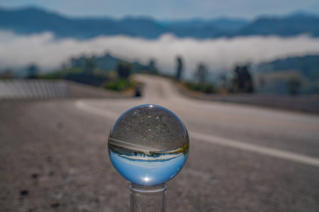 Sea of mist fills the valley as a curved road disappears into fog, captured through a glass sphere.の写真素材