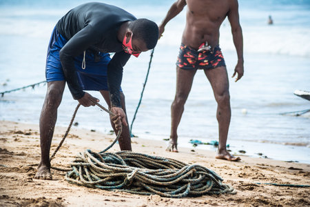 Salvador, Bahia, Brazil - May 23, 2021: Fisherman rolling fishing net to store in warehouse after fishing on Boca do Rio beach.のeditorial素材