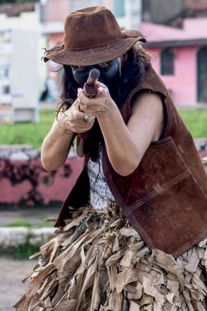 Caucasian woman with face made up in black color. She is wearing a pink hat and pointing a gun at...