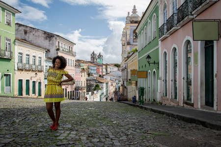 Model in a yellow dress smiling, looking and posing at the camera. In the background, the historical center of Pelourinho with its ancient architecture. Salvador Bahia Brazil.の写真素材