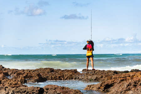 Salvador, Bahia, Brazil - October 10, 2021: Fisherman woman with fishing rod on the rocks fishing on the famous Rio Vermelho beach in Salvador, capital of Bahia, Brazilのeditorial素材