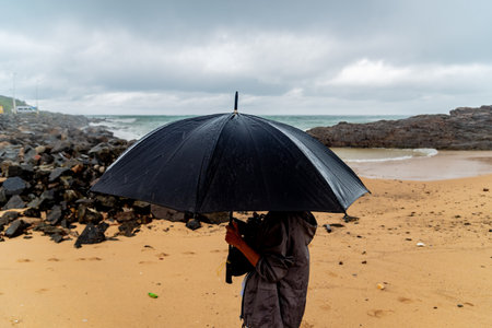 Salvador, Bahia, Brazil - November 15, 2021: A person holding a black umbrella against the sea in the background. Salvador, Bahia, Brazil.のeditorial素材