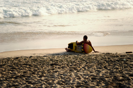 Salvador, Bahia, Brazil - September 26, 2021: Surfer sitting on the beach sand looking at the sea. Farol da Barra beach in Salvador, state of Bahia, Brazil.のeditorial素材
