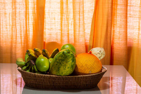 Fruits in a wooden basket on the glass table against orange background. Salvador, Bahia, Brazil.の写真素材