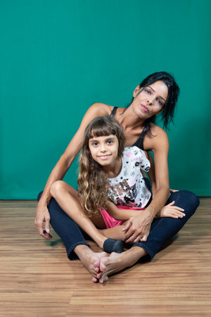 Portrait of mother and daughter sitting on wooden floor against dark green background. Salvador, Bahia, Brazil.の写真素材