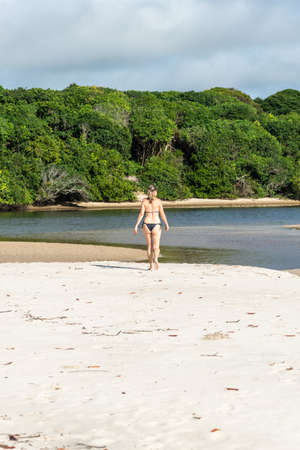 Young beautiful woman walking on the beach sand towards the trees and the mangrove. Praia do Guaibim, coast of the sea of Bahia, Brazilの写真素材