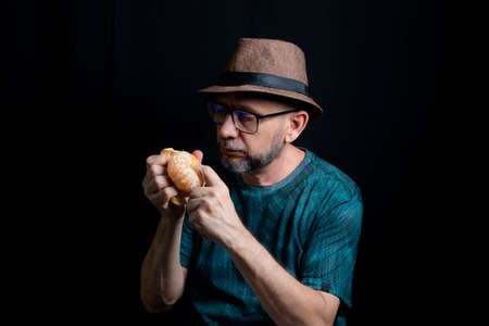 Man in glasses hat and beard peeling a tangerine against black background. Salvador, Bahia, Brazil.の写真素材