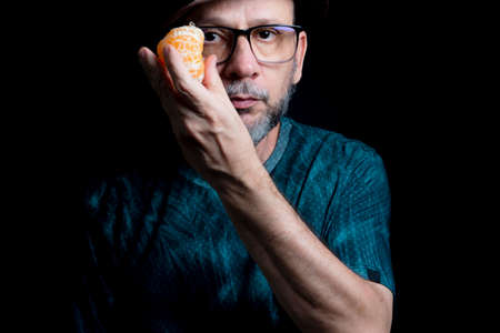 Man in glasses hat and beard holding a tangerine against black background. Salvador, Bahia, Brazil.の写真素材
