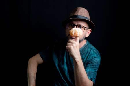 Man in glasses hat and beard holding a tangerine against black background. Salvador, Bahia, Brazil.の写真素材