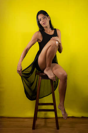 Portrait of model sitting on wooden bench holding black dress against yellow background in studio. Salvador, Bahia, Brazil.の写真素材