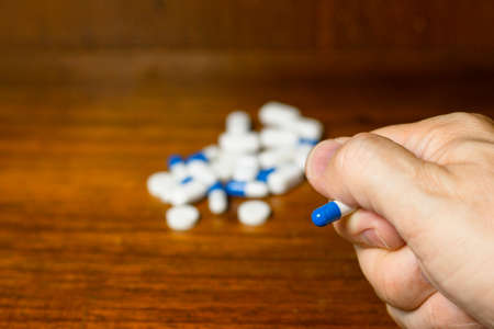 Simple pile of white, blue and yellow pills, medicines, pills stacked on brown background. Fingers...