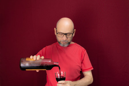 Young handsome man with beard, bald, red shirt wearing glasses holding in his hands a bottle of...