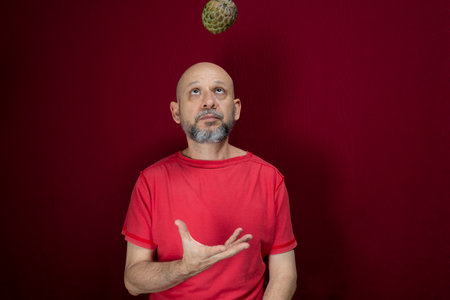 Young handsome man with beard, bald head and red shirt standing tossing pinecone fruit up against...