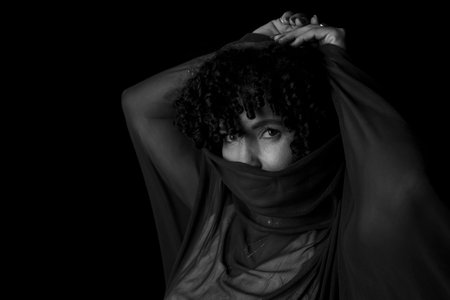 Close-up black and white portrait of woman with transparent cloth on her face against black...