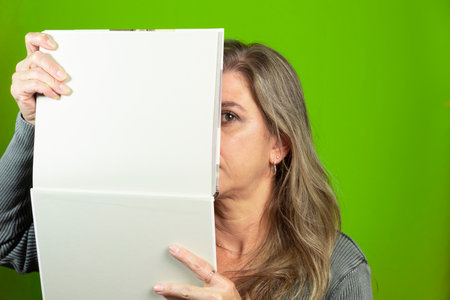 close-up portrait of a woman with her face behind a blank page against green background. Educational...