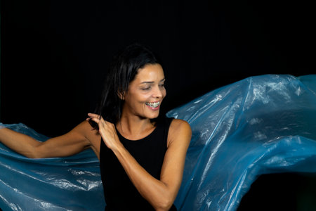 Portrait of woman with clear blue plastic behind her body against black background. She makes...