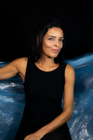 Portrait of woman with clear blue plastic behind her body against black background. She makes...