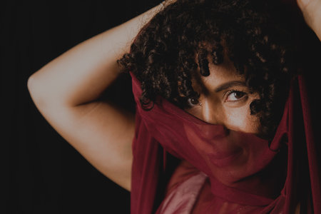 Portrait of beautiful woman with thin cloth of dark red color on her face against black background. Mature woman. Self-esteem concept.の写真素材