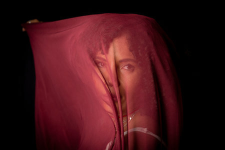 Portrait of beautiful woman with thin cloth of dark red color on her face against black background....