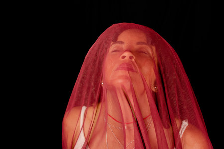 Portrait of beautiful woman with thin cloth of dark red color on her face against black background. Mature woman. Self-esteem concept.の写真素材