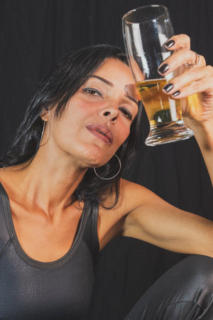 Portrait of young woman in black t-shirt drinking beer from glass cup. against black studio...