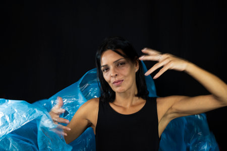 Portrait of woman with clear blue plastic behind her body against black background. She makes...