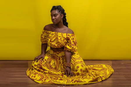 Young beautiful woman sitting on wooden floor, wearing colorful dress. Against yellow background.の写真素材