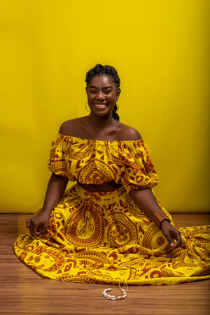 Young beautiful woman sitting on wooden floor, wearing colorful dress. Against yellow background.の写真素材