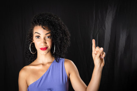 Portrait of young beautiful smiling woman pointing up. wearing blue clothes. Against black...