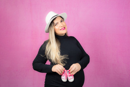 Pregnant woman in hat holding baby slippers. Isolated against pink background.