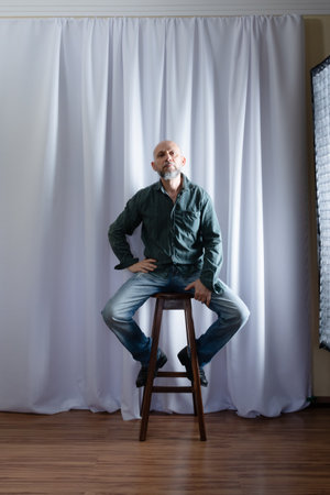 Studio portrait of a man sitting on a wooden stool. white curtain backgroundの写真素材