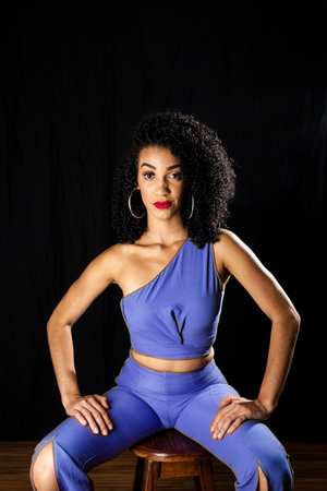 Young woman wearing blue outfit sitting on a wooden stool. Isolated black background.