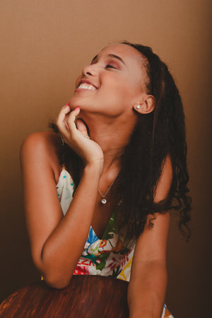 Portrait of pretty teenage girl leaning on a wooden stool with her hands on her chin. Against brown...