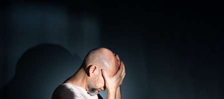 Sad man suffering from headache or migraine. Stressed guy with painful facial expression feeling terrible weakness or depression. Isolated on dark background.の写真素材