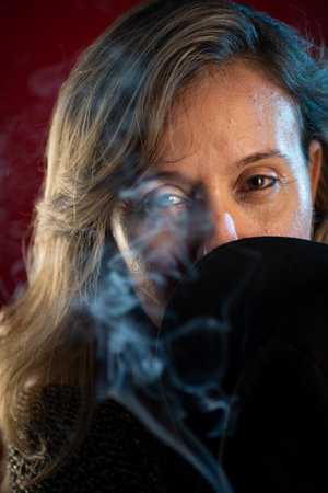 Attractive, confident woman holding a black beret in front of her face. Incense smoke all around. Isolated on a red background.の写真素材