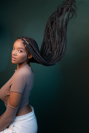 Beautiful young woman, sitting sideways, with braids of her hair flying. Isolated on dark cyan background.の写真素材