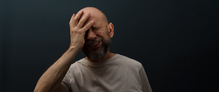 Sad man suffering from headache or migraine. Stressed guy with painful facial expression feeling terrible weakness or depression. Isolated on dark background.の写真素材