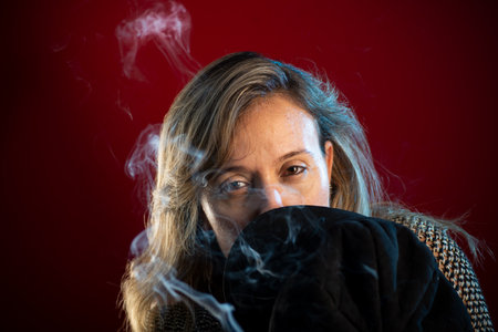 Attractive, confident woman holding a black beret in front of her face. Incense smoke all around. Isolated on a red background.の写真素材