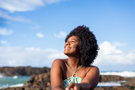 Portrait of a smiling woman wearing blue sitting on the rock of a beach looking to the side. Blue sky in the background. Rio Vermelho Beach, Salvador, Brazil.の写真素材