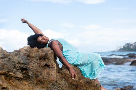Portrait of a beautiful woman with black power hair lying on the rocks of a beach wearing light blue...