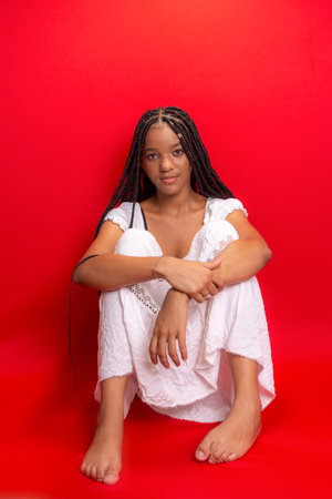 Beautiful young black woman sitting and serene wearing white outfit with long braids in her hair. Isolated on red backgroundの写真素材